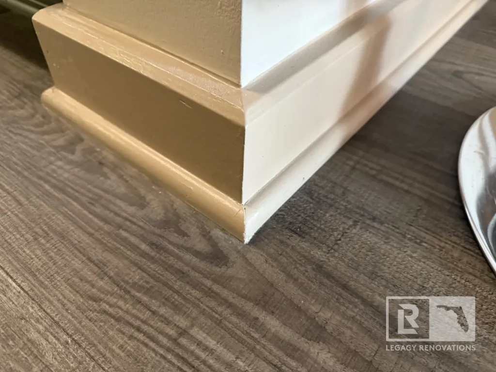 Close-up corner view of beige painted baseboard trim and mitered shoe molding resting along dark wood-grain vinyl plank flooring.