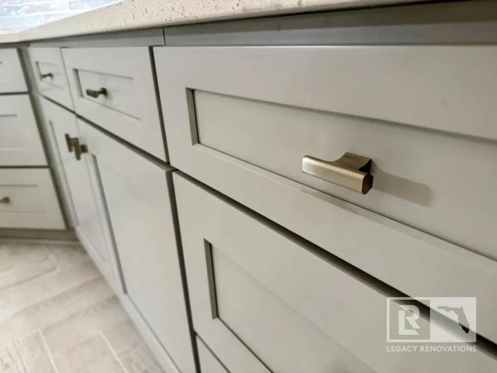 Close-up view of brushed nickel metal pulls attached to light grey shaker drawer fronts below a white quartz countertop with light grey wood-look floor tile.