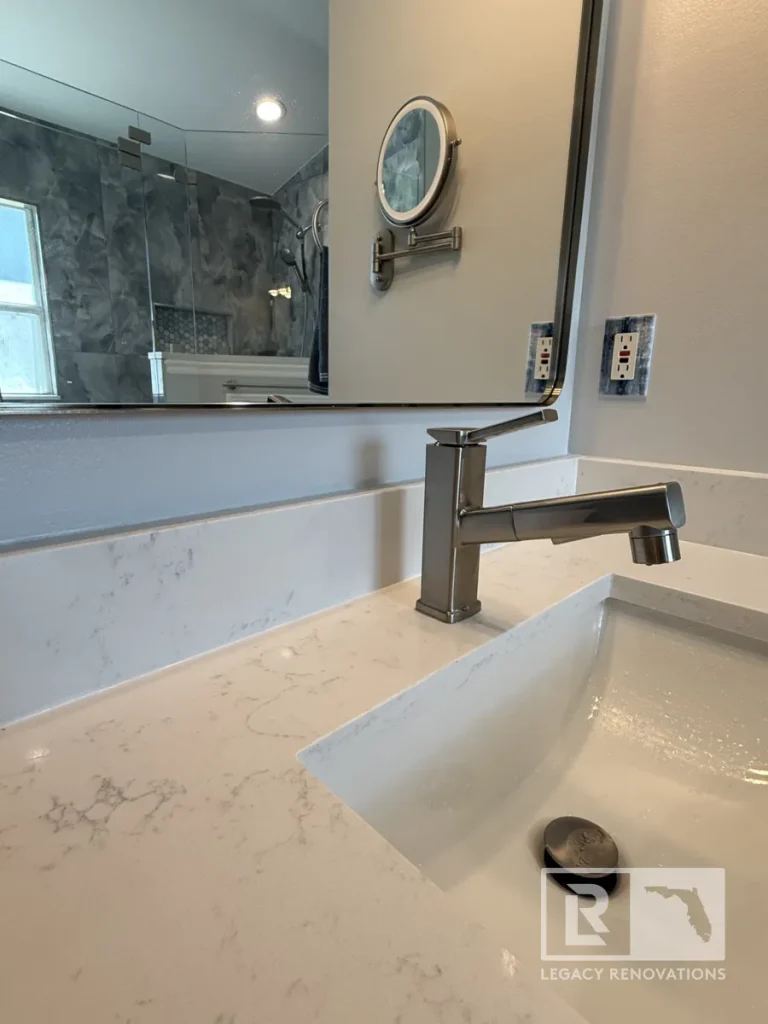 Angular brushed metal sink spout installed on a white marble-veined quartz countertop above a rectangular undermount basin with mirror reflection showing a dark grey tiled shower.