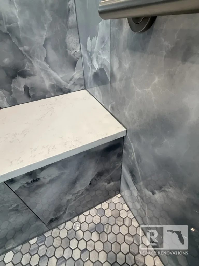 Built-in corner shower bench clad in dark grey marbled tile topped with a white quartz slab, positioned above small hexagonal floor tiles with a metal handrail visible at the top.