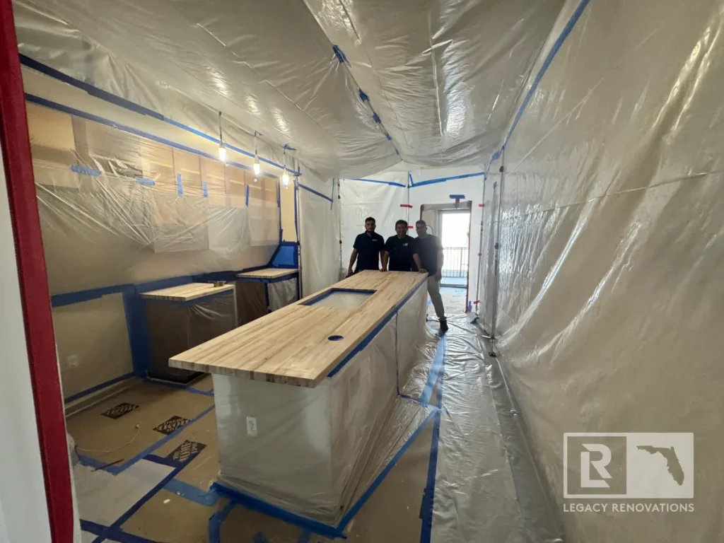 Three contractors standing behind an unfinished wood butcher block island inside a kitchen completely covered in clear plastic dust barriers taped with blue tape.