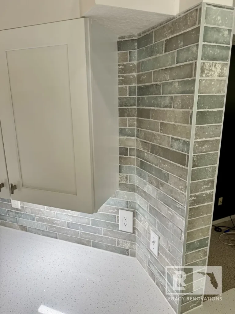 Vertical view of a wall corner wrapped in distressed green and grey brick-style ceramic tiles with white Schluter trim edging next to a upper cabinet and two white electrical outlets.