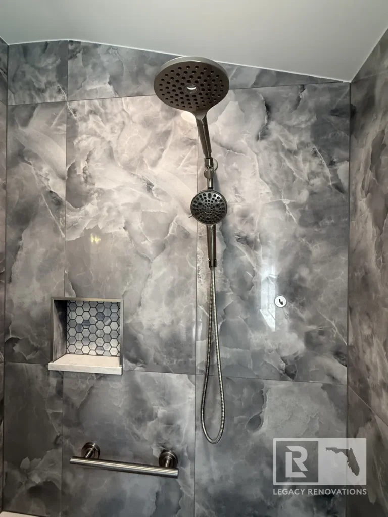 Brushed metal dual showerhead assembly with attached flexible hose mounted on cloud-patterned grey wall tiles above a horizontal safety grab bar and beside a hexagonal tile niche.