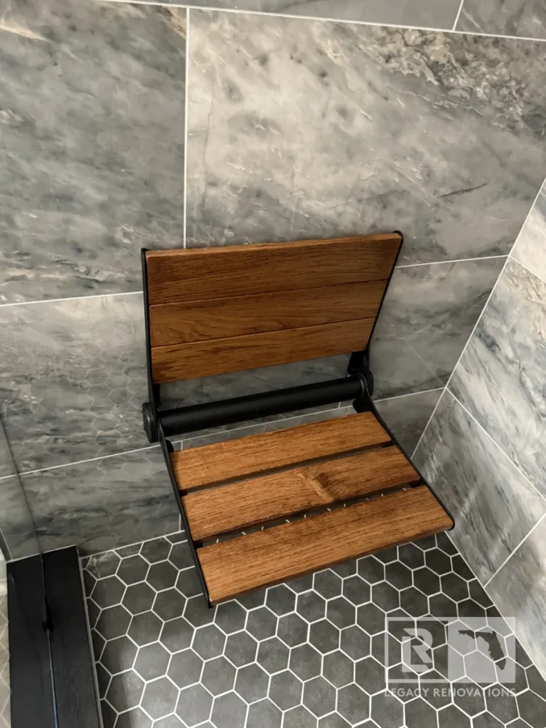 Wall-mounted folding teak shower seat with black frame above dark hexagon floor tile in Jacksonville, FL
