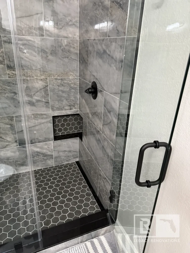 Glass-enclosed shower with gray veined wall tile, black hexagon floor, recessed niche, and black handle in Jacksonville, FL