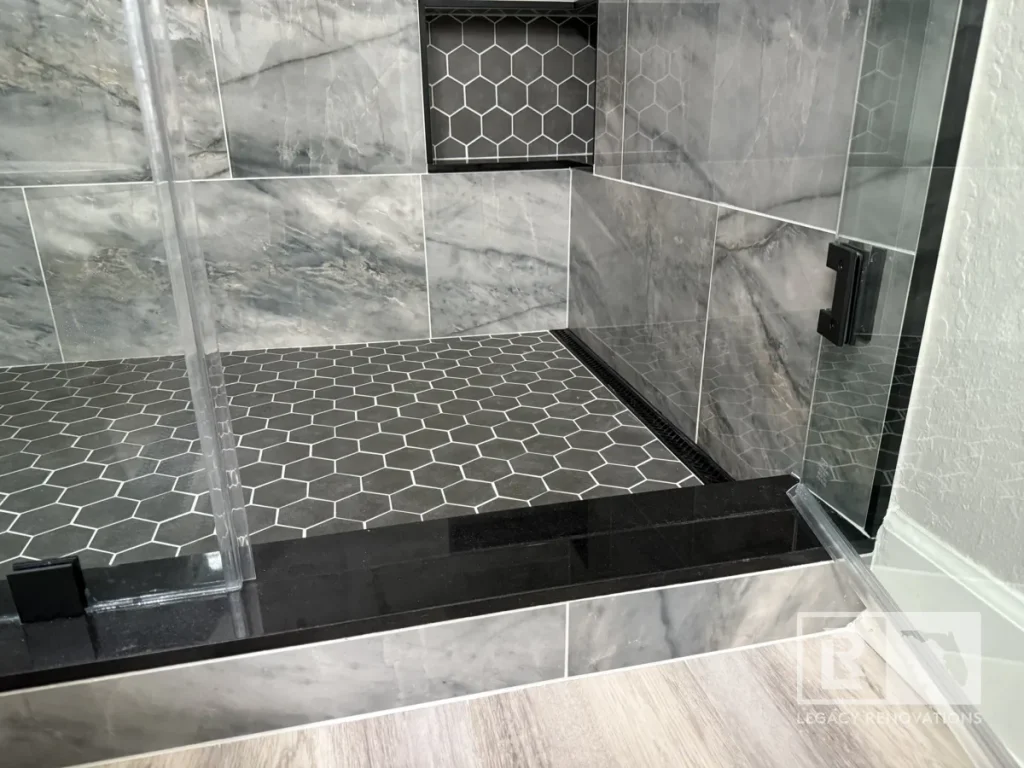 Close-up of black hexagon shower floor tile, gray wall tile, recessed niche, and glass enclosure in Jacksonville, FL