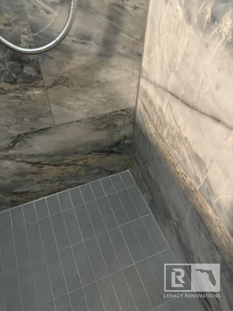 Close-up corner view showing dark grey rectangular mosaic floor tile meeting veined wall tiles, with a metallic flexible shower hose visible above.