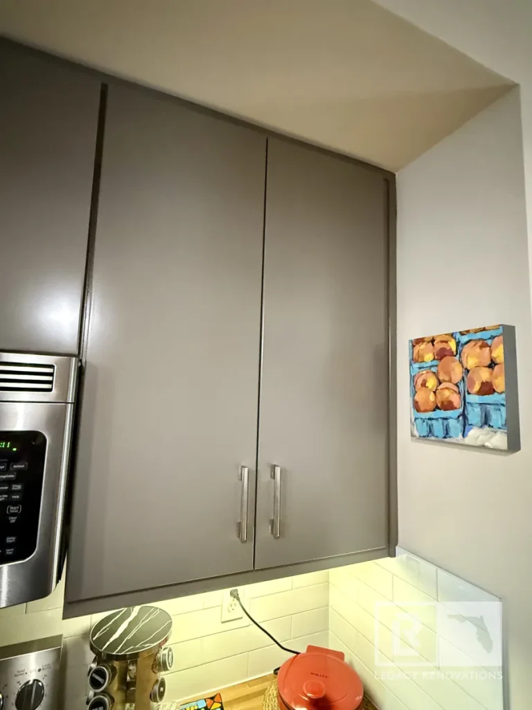 Flat-panel grey upper kitchen cabinet doors with brushed silver handles positioned over white subway tile backsplash, under-cabinet lighting, and a small fruit painting on the adjacent wall.