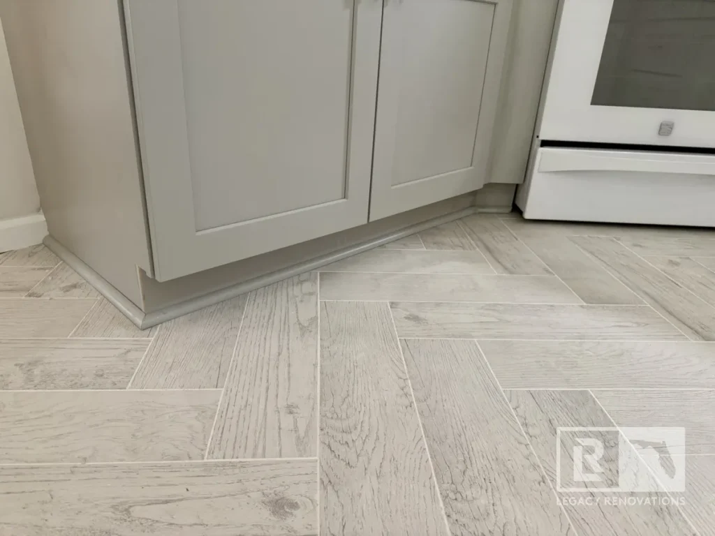 Ground level perspective showing ash grey wood-look ceramic tiles arranged in a herringbone pattern meeting the baseboard quarter round of a lower cabinet frame.
