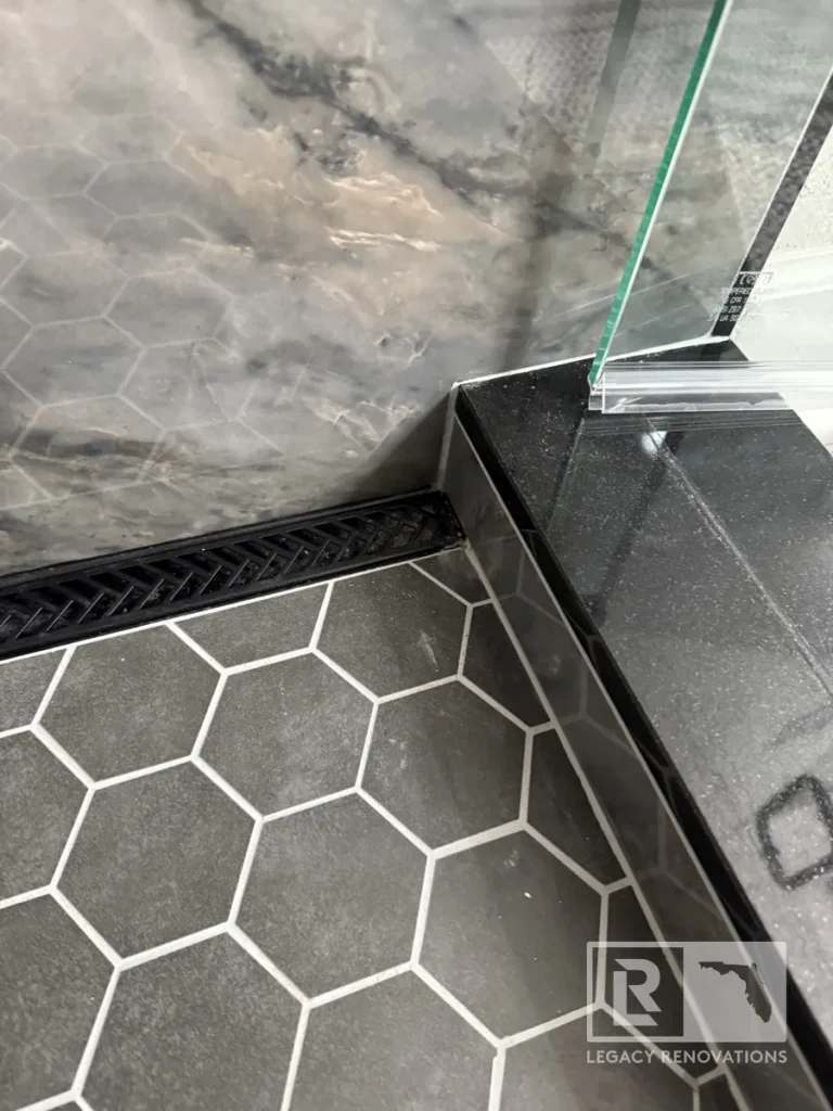Close-up of dark gray hexagon shower floor tiles, black linear drain, gray veined wall tile, and glass panel in Jacksonville, FL