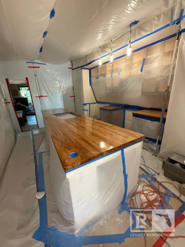 Kitchen under construction completely sealed in white plastic sheeting with blue painter tape, featuring a glossy wood butcher block island with sink cutout and hanging pendant light bulbs.