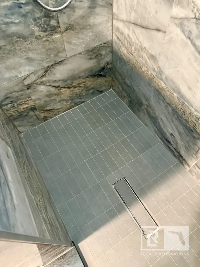 High-angle view of a grey rectangular mosaic tile shower pan featuring a long metallic linear trench drain and marbled wall panels.