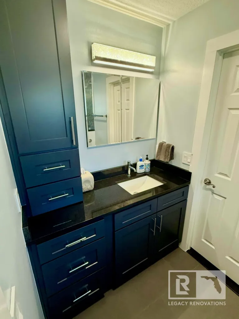 Full view of a dark blue vanity cabinet featuring a black granite top, undermount sink, mirrored medicine cabinet, horizontal LED light fixture, side linen tower, and white interior door.