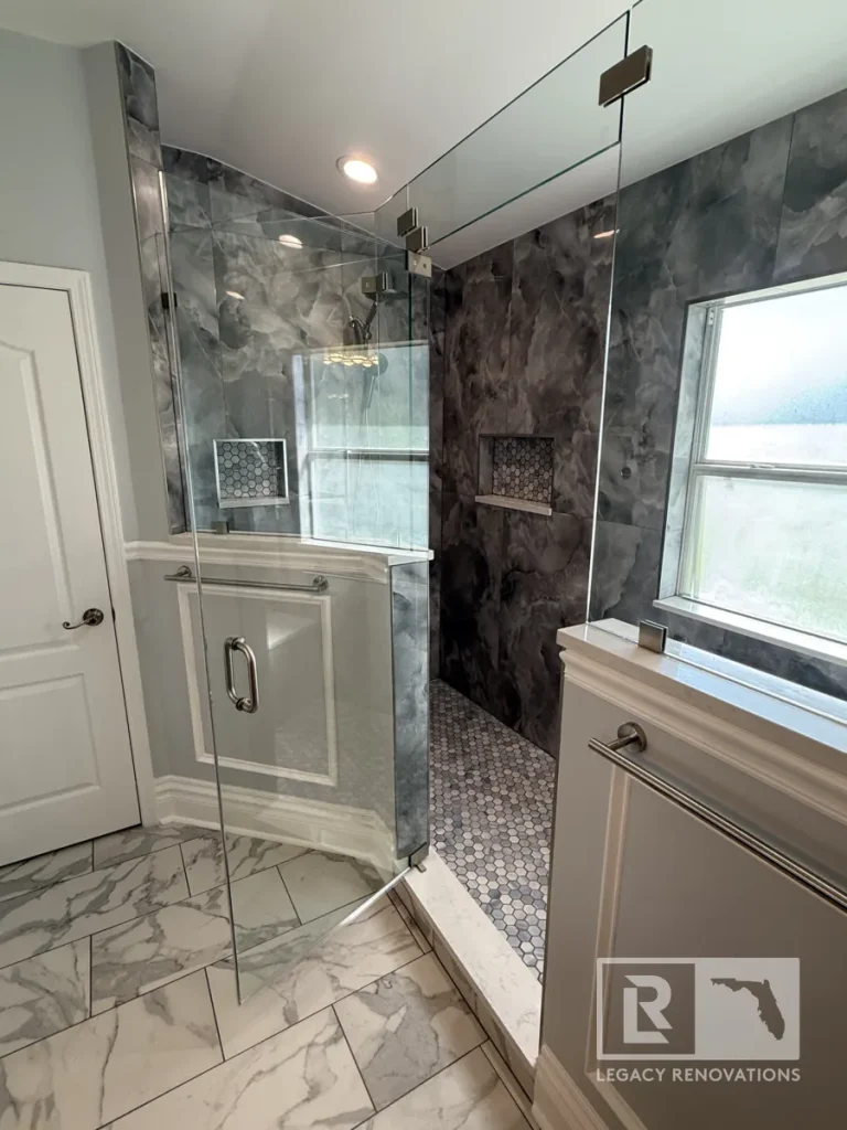 Open frameless glass shower door revealing a tiled stall with dual niches, a window, grey hexagon floor tiles, and a white interior door.