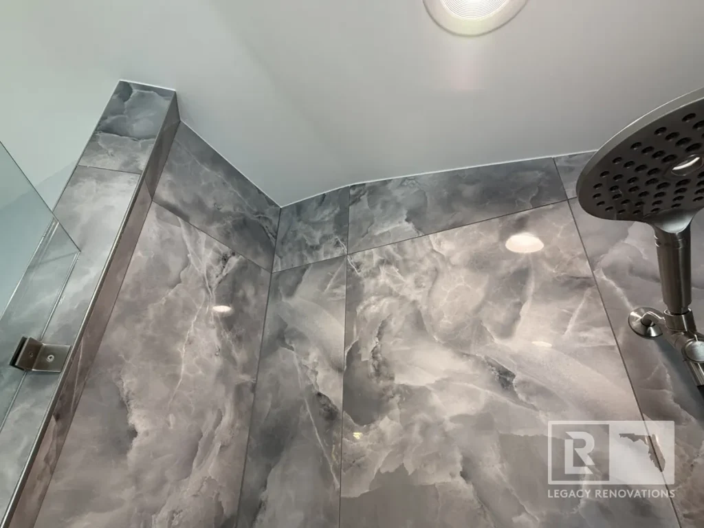 Low-angle view looking up at a circular brushed nickel shower head, recessed ceiling light, and glossy slate-grey marbled wall tile.