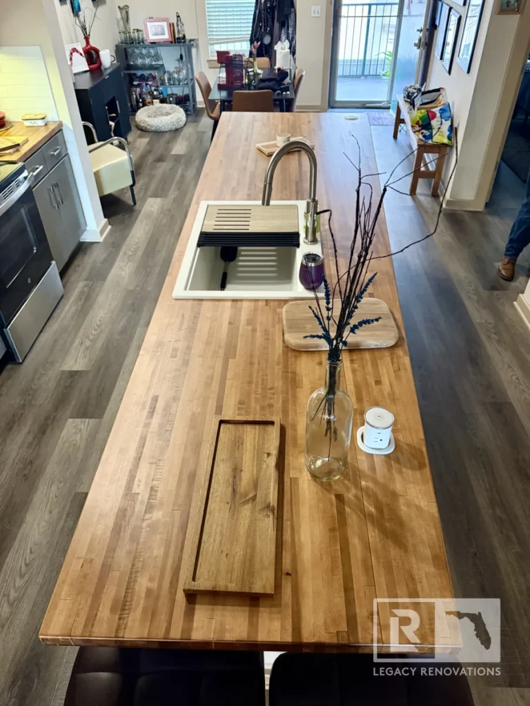 High-angle full view looking down the length of a butcher block island featuring a clear vase with dark stems, a wooden serving tray, an undermount white sink, and dark bar stool seating.