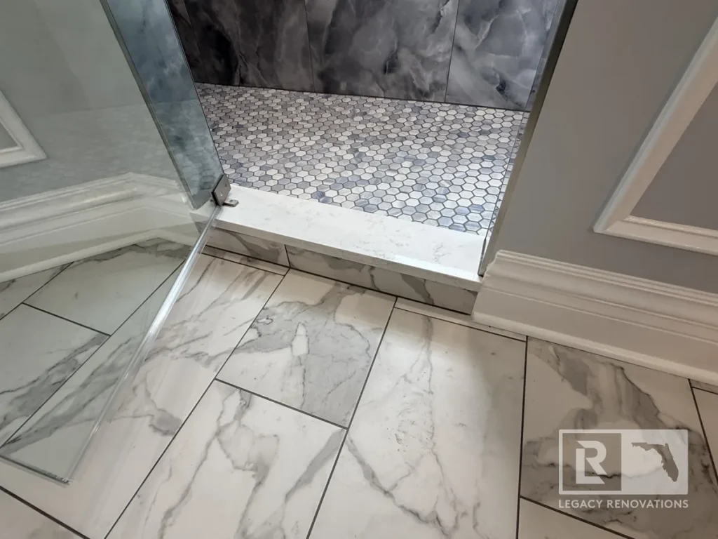 Low-angle shot showing a white quartz shower threshold curb separating white-and-grey marble-patterned floor tiles from small grey hexagon shower pan tiles next to an open glass door.