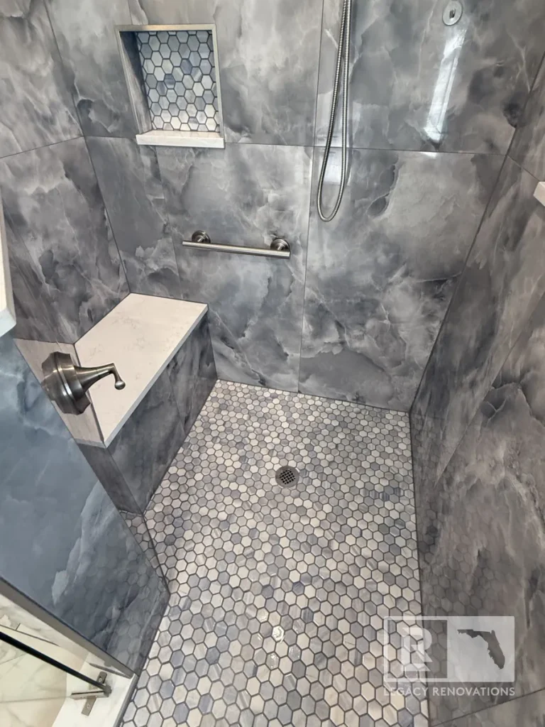 Top-down view inside a shower enclosure showing a metallic water control handle mounted on the side wall near a quartz-topped bench, a metallic hose line, and a circular floor drain set into grey hexagon tiles.