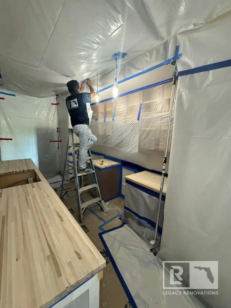 Worker on a stepladder taping plastic sheeting near temporary lights along the wall, with unfinished butcher block counters and tension support poles visible below.