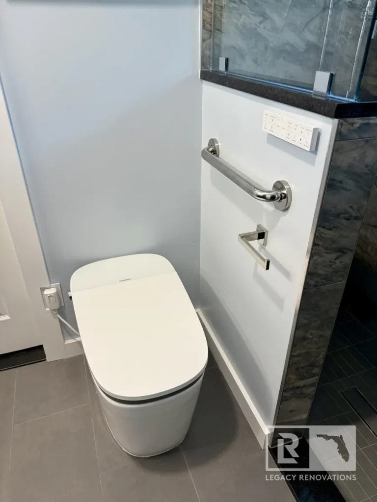 White tankless bidet toilet positioned on grey floor tile next to a half wall equipped with a wall-mounted remote control panel, horizontal grab bar, and toilet paper holder.