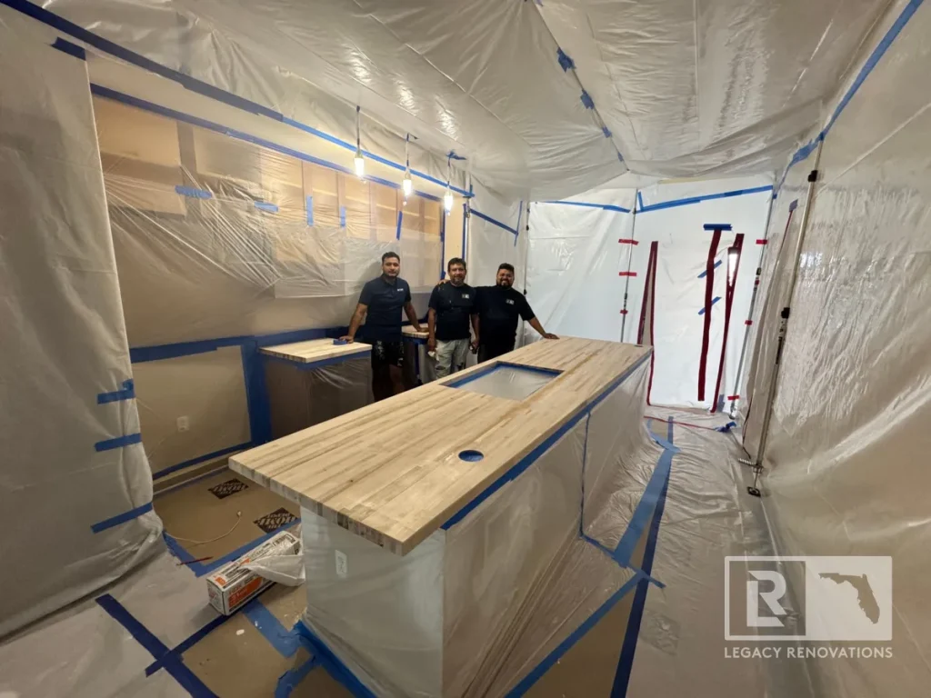Three men in dark shirts stand side-by-side behind an unfinished butcher block kitchen island surrounded by full plastic wall and ceiling protective sheeting.
