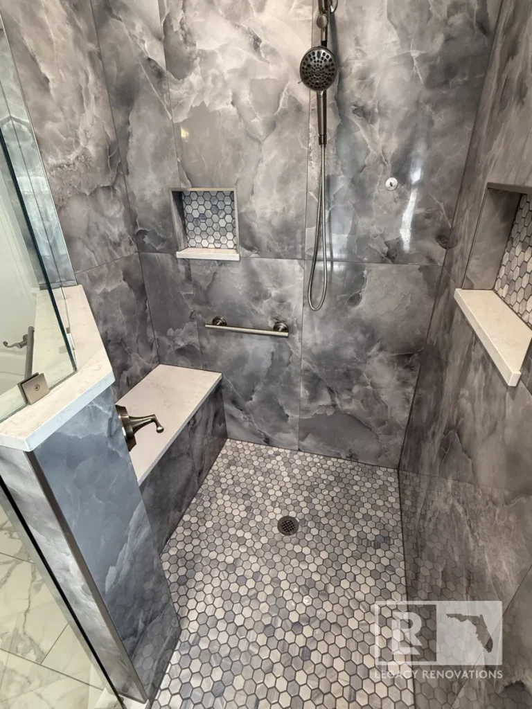 Elevated view looking down into a tiled shower stall showing a central round drain set in honeycomb mosaic floor tile, two recessed wall alcoves, a bench, and a handheld wand sprayer.