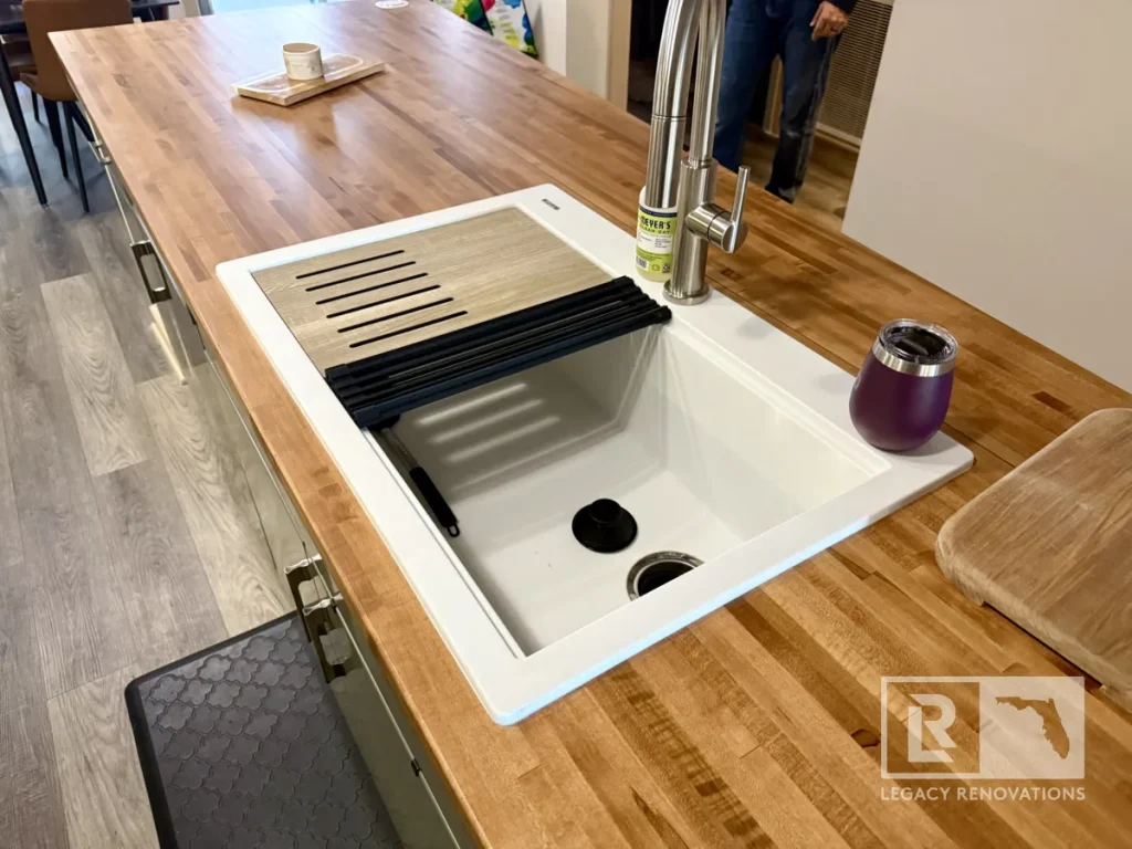 Angled top-down view of a white rectangular workstation basin drop-in sink with slotted cutting board, black drying rack insert, brushed nickel faucet, and purple tumbler on a butcher block island.