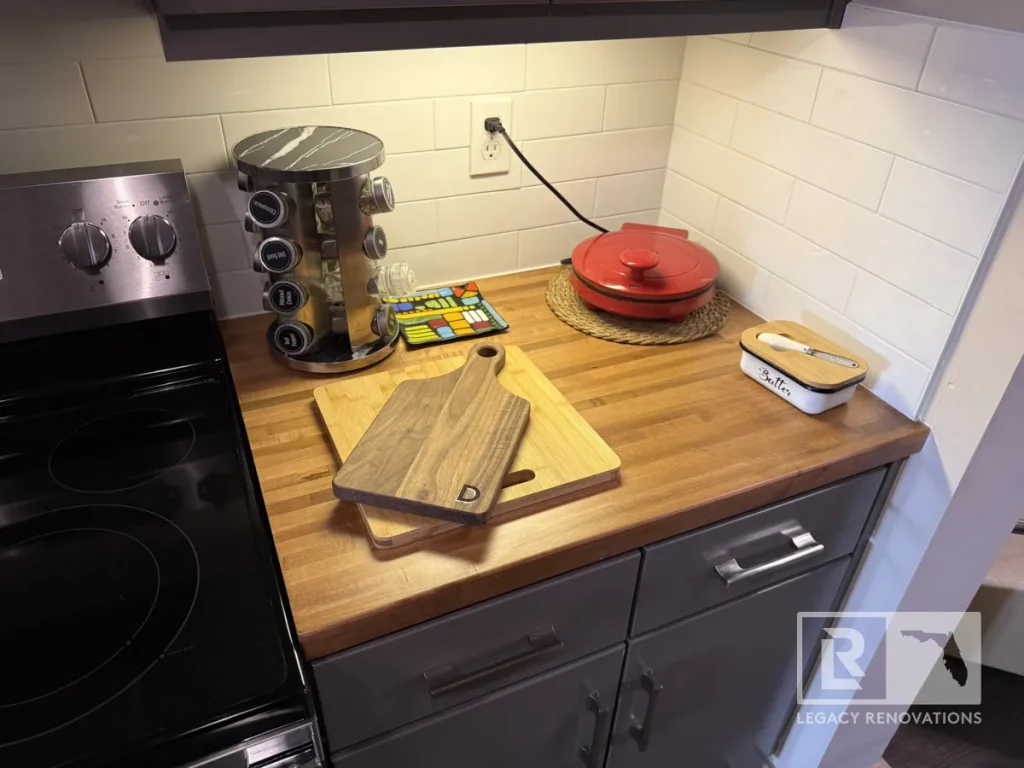 Kitchen counter corner featuring stacked wooden cutting boards, revolving metal spice rack, red round appliance, butter dish, white subway tile backsplash, and electric glass-top range.