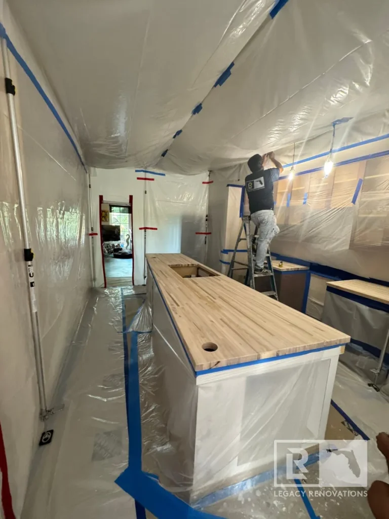 Tradesman on a step ladder applying blue painter tape to plastic ceiling sheeting inside a prepped kitchen with raw butcher block countertops and extension poles.