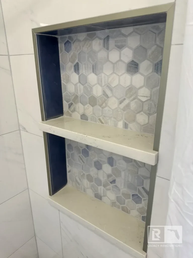 Angled view of a custom recessed double shower niche with gray hexagon mosaic tile, quartz shelves, and marble-look wall tile in a Jacksonville, FL bathroom remodel.