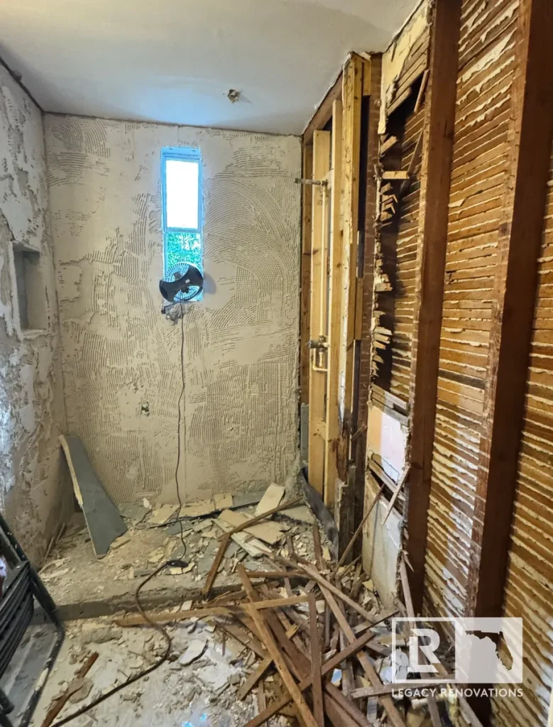 Bathroom renovation demolition stage with exposed wood lath framing, removed tile mortar bed, debris on the floor, and a small transom window.
