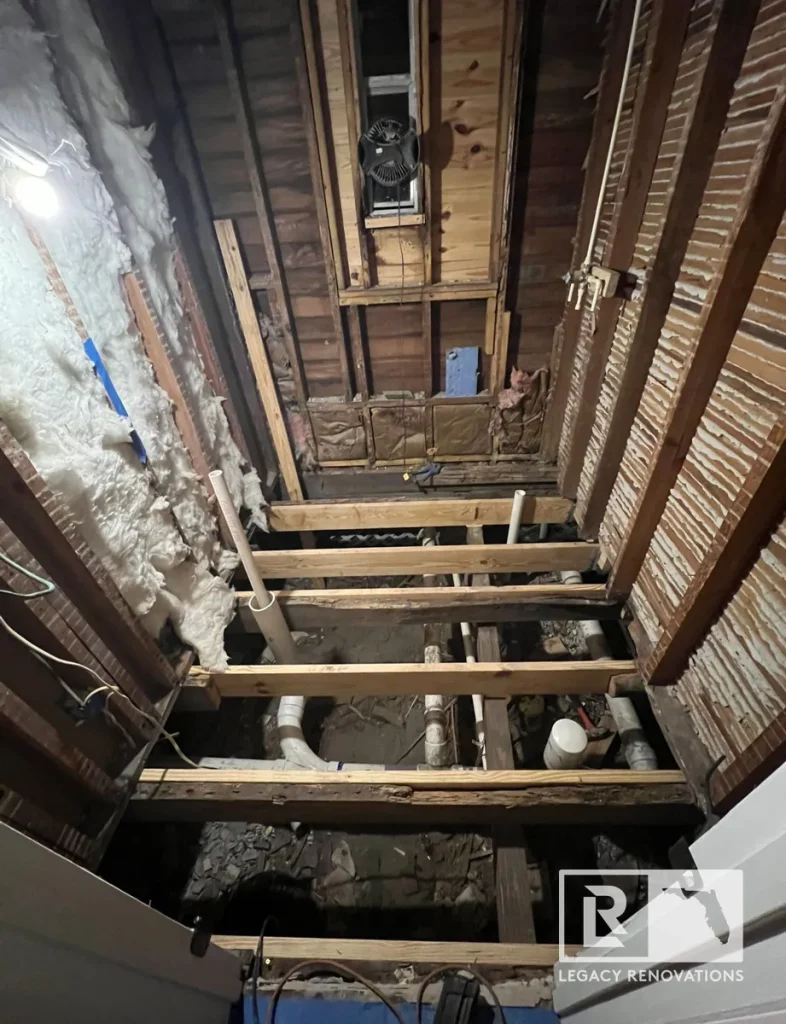 Gutted bathroom during renovation showing exposed floor joists, rough plumbing pipes, wall framing, and insulation.