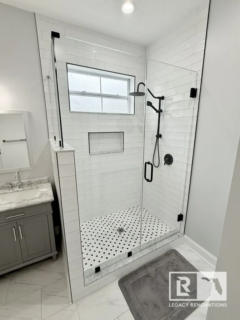 Close-up of a contemporary frameless glass walk-in shower featuring white subway tile, matte black rainfall shower fixtures, recessed wall niche, and penny tile flooring.