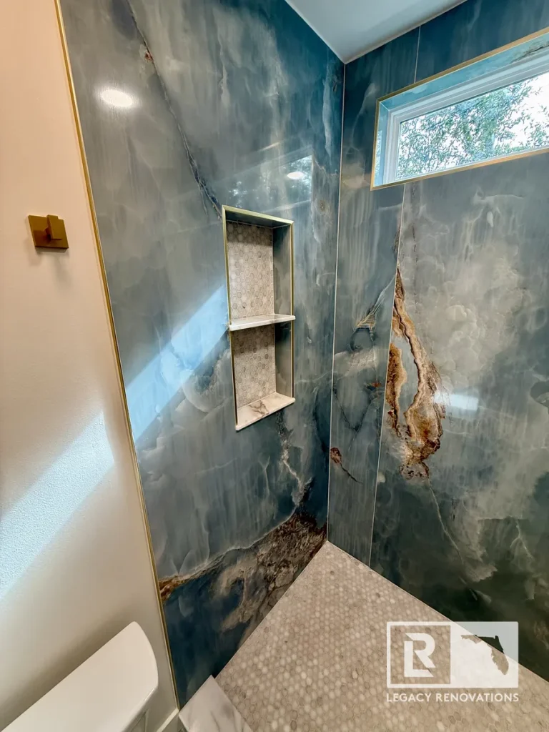 Custom walk-in shower with blue onyx-look porcelain slab walls, double recessed niche, mosaic tile floor, and transom window in a Jacksonville, FL bathroom remodel