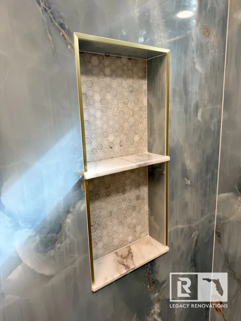 Double recessed shower niche with brushed brass trim, marble shelves, hexagon mosaic tile, and blue onyx-look porcelain walls in a Jacksonville, FL bathroom remodel