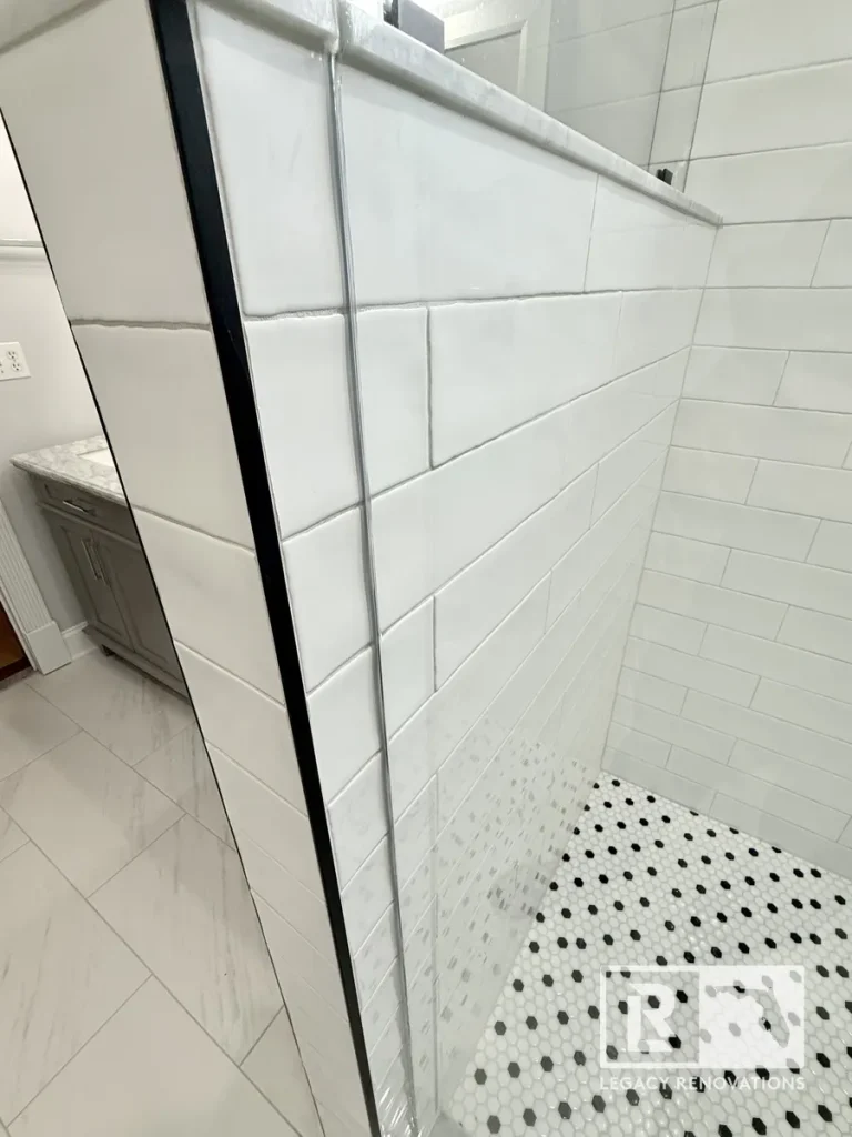 Frameless glass shower enclosure with glossy white subway tile walls, marble curb, and black-and-white penny tile flooring in a modern bathroom.