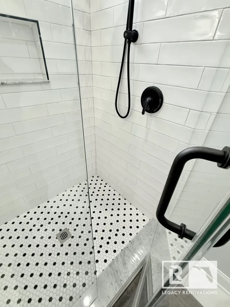 Frameless glass walk-in shower with white subway tile walls, matte black shower fixtures, marble curb, and black-and-white penny tile flooring.