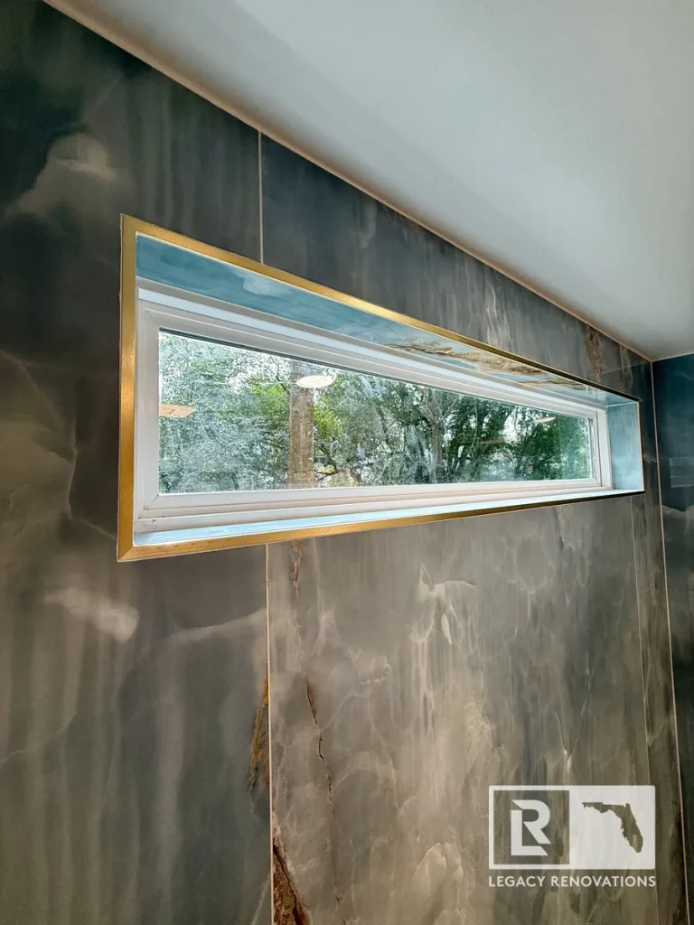 Horizontal shower window surrounded by blue onyx-look porcelain slab walls with brushed brass trim in a luxury Jacksonville, FL bathroom remodel