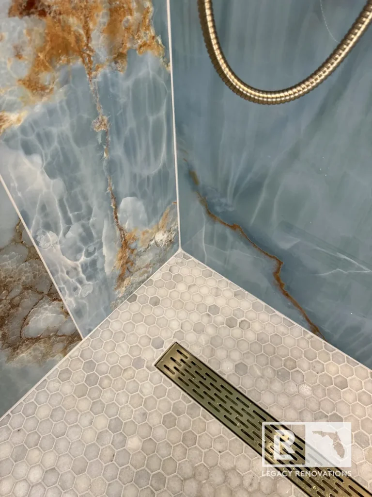 Close-up of a brushed bronze linear shower drain with white hexagon mosaic tile floor and blue onyx-look porcelain walls in a Jacksonville, FL bathroom remodel