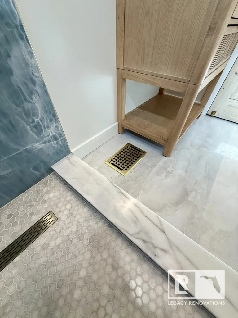 Close-up of a marble shower threshold with a linear drain, hexagon mosaic tile floor, brushed brass floor register, and wood vanity in a Jacksonville, FL bathroom remodel