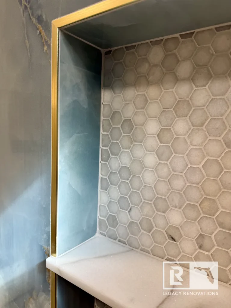 Custom recessed shower niche with white hexagon mosaic tile, quartz shelf, brushed brass trim, and blue onyx-look porcelain walls in a Jacksonville, FL bathroom remodel