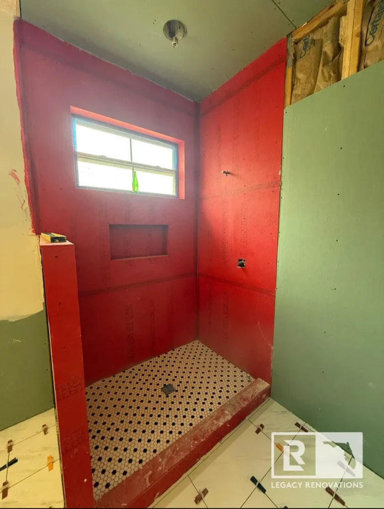 Walk-in shower under construction featuring a red waterproofing membrane, recessed shower niche, marble curb, and black-and-white penny tile shower floor.