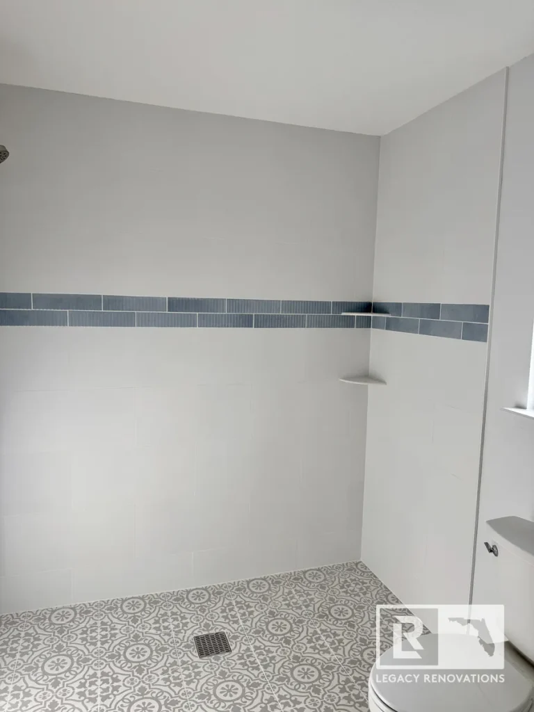 Finished walk-in shower with large-format wall tile, blue accent tile, built-in corner shelf, and patterned porcelain floor in a bathroom remodel in Jacksonville, FL.