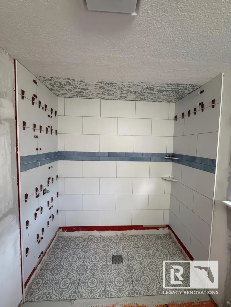 In-progress tile shower installation in Jacksonville, FL featuring white subway wall tiles, a blue accent band, tile leveling wedges, and patterned floor mosaic.
