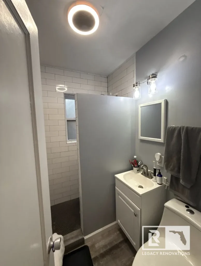 Completed small bathroom remodel with a white vanity, walk-in subway tile shower, modern lighting, gray walls, and wood-look flooring in Jacksonville, FL.