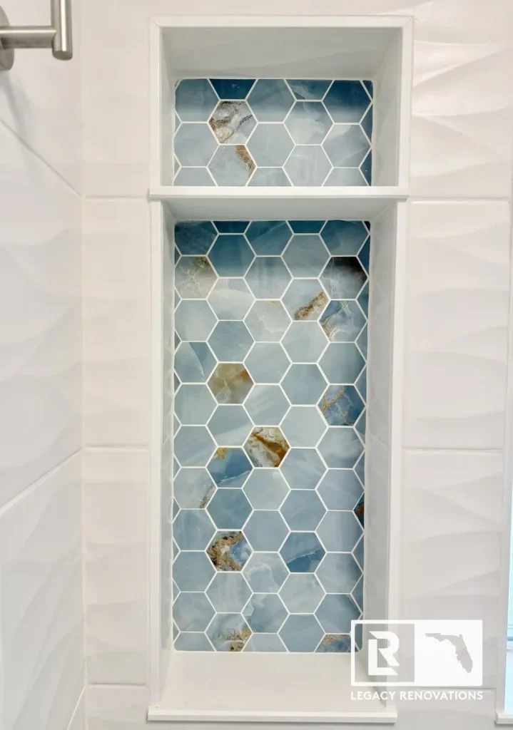 A finished tall recessed shower niche with two storage shelves, backed with blue onyx-style hexagon mosaic tile and framed by white textured wave wall tiles by Legacy Renovations in Jacksonville, FL.