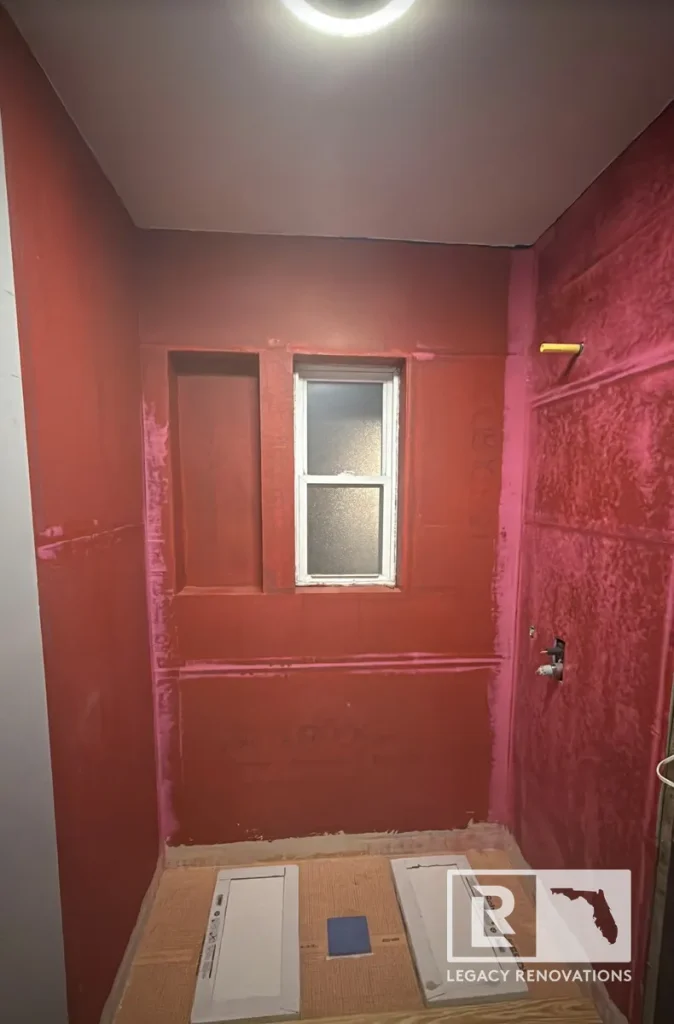 Shower waterproofing in progress with red waterproofing membrane, recessed niche, and frosted window during a bathroom remodel in Jacksonville, FL.