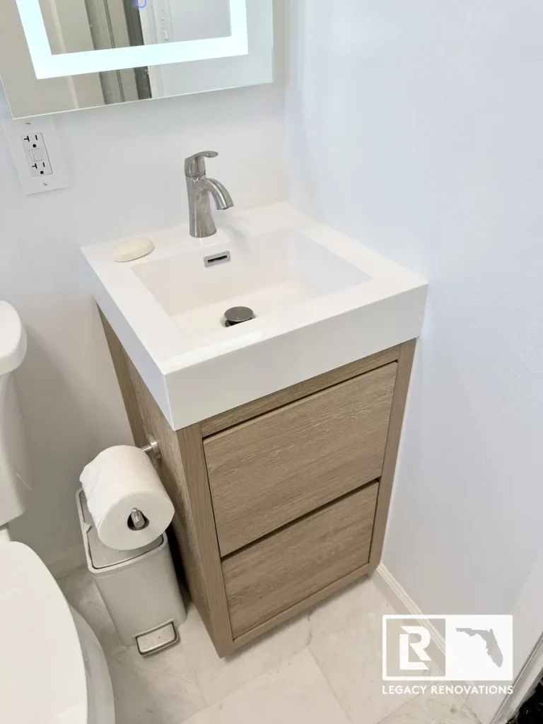 A finished small bathroom corner featuring a light wood two-drawer vanity, integrated white square sink, brushed nickel faucet, and a backlit LED mirror by Legacy Renovations in Jacksonville, FL.