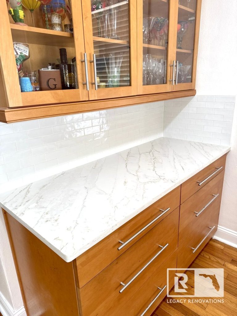 Kitchen remodel in Jacksonville, FL featuring a built-in buffet with natural wood cabinets, glass-front upper cabinets, quartz countertops, white subway tile backsplash, and modern hardware.