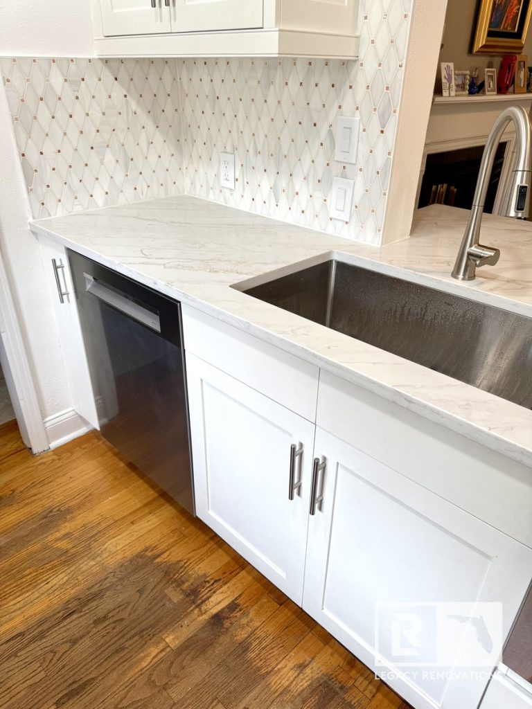 Kitchen remodel in Jacksonville, FL featuring white shaker-style cabinets, quartz countertops, a stainless steel farmhouse sink, geometric tile backsplash, stainless dishwasher, and hardwood flooring.